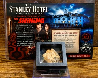 May include: A rectangular display case containing a rough, tan-colored architectural stone fragment. Behind the case is a card featuring The Stanley Hotel, the text "Filming Location of The Shining," and a certificate of authenticity.