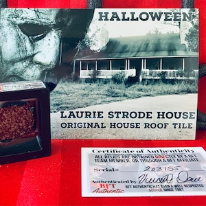 Op de afbeelding: Een ingelijst stuk dakpan van het Laurie Strode huis, met een certificaat van echtheid. De afbeelding bevat een foto van het huis en het woord "HALLOWEEN". Het serienummer is 203155.