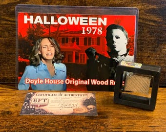 Może przedstawiać: Prostokątna karta wystawowa z napisem HALLOWEEN 1978 oraz Doyle House Original Wood Relic, przedstawiająca scenę filmową z osobą i zamaskowaną postacią. Zawiera certyfikat autentyczności oraz kawałek drewna umieszczony w czarnej ramce.