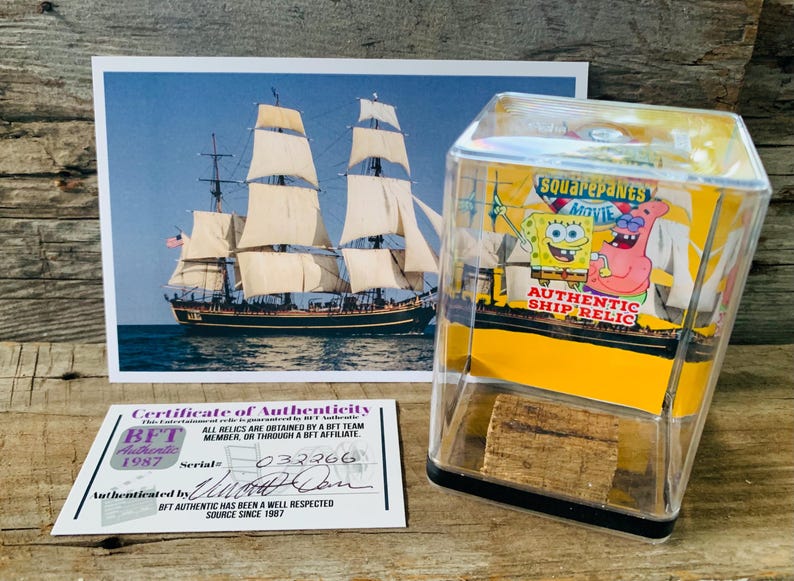 May include: A clear plastic display case with a yellow base and a black trim. The case contains a small piece of wood and a printed image of Spongebob Squarepants and Patrick Star from the movie "Squarepants Movie". The text "Authentic Ship Relic" is printed on the case.