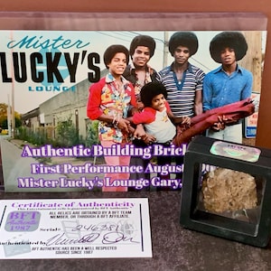 Puede incluir: Imagen que muestra una fotografía enmarcada con el texto "Mister Lucky's Lounge" y "Authentic Building Brick". También se ve un certificado de autenticidad y una pequeña vitrina con una roca.