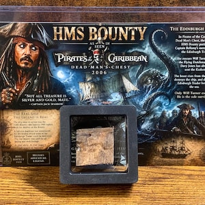 May include: A rectangular display card featuring Captain Jack Sparrow and the HMS Bounty from Pirates of the Caribbean: Dead Man's Chest, with a small, square, weathered wooden fragment encased in a black frame mounted on the front of the card.