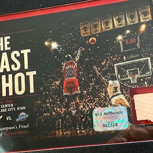 May include: A framed commemorative plaque titled The Last Shot featuring Michael Jordan in a red jersey taking a shot against the Utah Jazz on June 14, 1998. The image includes a holographic BET Authentic sticker and a small piece of light wood.