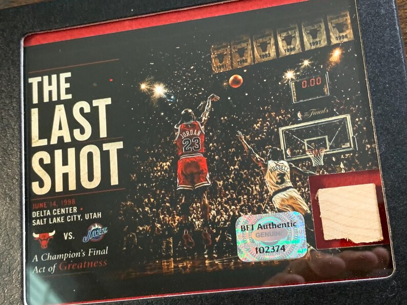 May include: A framed commemorative plaque titled The Last Shot featuring Michael Jordan in a red jersey taking a shot against the Utah Jazz on June 14, 1998. The image includes a holographic BET Authentic sticker and a small piece of light wood.