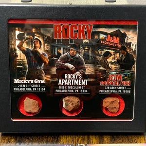 May include: A black framed display case featuring a Rocky movie collage with three circular cutouts containing small, rough, reddish-brown stone fragments. The print includes images of Micky's Gym, Rocky's Apartment, and J&M Tropical Fish with addresses.
