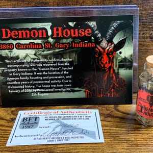 May include: A clear glass vial with a cork stopper containing soil, accompanied by a rectangular display card titled Demon House 3860 Carolina St. Gary Indiana featuring a goat illustration and a white Certificate of Authenticity card with BFT branding.