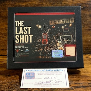May include: A black framed display featuring a photograph of Michael Jordan taking The Last Shot in 1998. The display includes a small piece of wood, a holographic BFT Authentic sticker, and a separate white Certificate of Authenticity card.