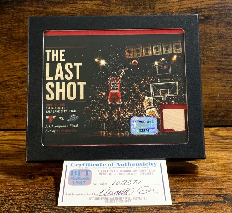 May include: A black framed display featuring a photograph of Michael Jordan taking The Last Shot in 1998. The display includes a small piece of wood, a holographic BFT Authentic sticker, and a separate white Certificate of Authenticity card.