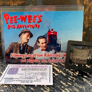 May include: A framed collectible from the movie "Pee-Wee's Big Adventure" featuring a scene with Pee-Wee Herman and a train. Includes a certificate of authenticity and a small, framed relic. The text reads "Filming Location Railroad".