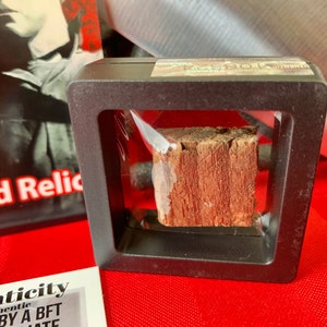 Pu&ograve; includere: Un piccolo pezzo di legno racchiuso in una teca di plastica nera con un coperchio di plastica trasparente. Il legno &egrave; di colore marrone rossiccio e ha una consistenza ruvida. La teca ha un'etichetta che dice "Reliquia di legno autentica".