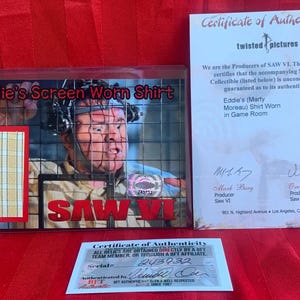 May include: A framed display of a screen-worn shirt from the film Saw VI. The shirt is white with a red and black "SAW VI" logo. The display includes a certificate of authenticity signed by the producers, Mark Burg and Oren Koules.