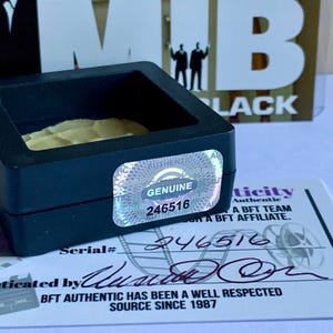 Puede incluir: Una vitrina negra con una pegatina hologr&aacute;fica "GENUINE" y el n&uacute;mero "246516". Se ve un certificado de autenticidad, con el texto "BFT AUTHENTIC HAS BEEN A WELL RESPECTED SOURCE SINCE 1987". El fondo presenta el texto "MIB BLACK".