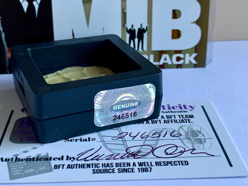 Puede incluir: Una vitrina negra con una pegatina hologr&aacute;fica "GENUINE" y el n&uacute;mero "246516". Se ve un certificado de autenticidad, con el texto "BFT AUTHENTIC HAS BEEN A WELL RESPECTED SOURCE SINCE 1987". El fondo presenta el texto "MIB BLACK".
