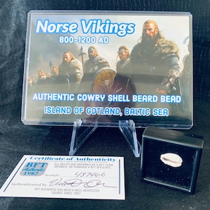 May include: A certificate of authenticity for a cowry shell beard bead from the island of Gotland in the Baltic Sea. The certificate is from BFT Authentic, a company that has been in business since 1987. The certificate has a serial number of 487466 and a signature. The bead is in a small black box.