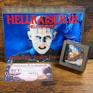 Puede incluir: Una exhibición de Hellraiser III Hell on Earth que incluye un póster de película, un pequeño fragmento marrón en un marco cuadrado negro y un Certificado de Autenticidad con el texto BFT Authentic y el número de serie 132939 en el documento.