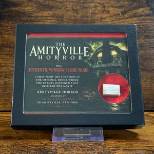 May include: A black display box containing a small piece of white wood labeled as authentic window frame wood from The Amityville Horror house. The text on the display details the location at 112 Ocean Avenue in Amityville, New York.