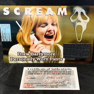 May include: A Scream movie prop, a pair of black pants worn by Drew Barrymore in the film. The pants are displayed on a card with a photo of Barrymore in the film and the Scream Ghostface mask. The card has a certificate of authenticity from BFT Authentic.