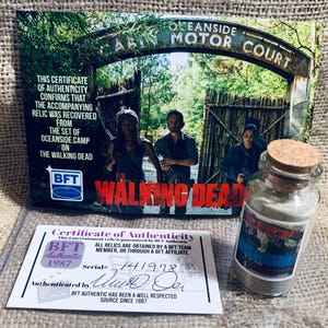 Puede incluir: Un certificado de autenticidad para una reliquia de The Walking Dead. El certificado es de BFT Authentic y tiene el número de serie 141978. El certificado está autenticado por "Ward Oer". El certificado va acompañado de un pequeño frasco de vidrio con tapa de corcho. El frasco contiene una pequeña cantidad de tierra. El frasco tiene una etiqueta que dice "The Walking Dead".