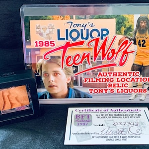 May include: A framed photograph of a liquor store called "Tony's Liquors" with the text "1985 Teen Wolf Authentic Filming Location Relic" and a certificate of authenticity with the text "BFT Authentic 1987 Serial: 032342 Authenticated by ... BET Authentic has been a well respected source since 1987."