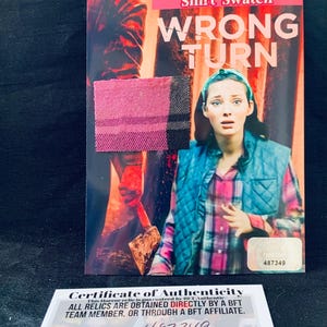 May include: A "Wrong Turn" movie prop featuring a shirt swatch and a certificate of authenticity. The main image shows a woman in a plaid shirt and a blue vest. The certificate has the serial number 487349.