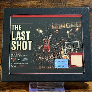 May include: A black framed collectible display featuring a photograph of Michael Jordan taking The Last Shot in 1998. The print includes the text The Last Shot, June 14, 1998, Delta Center, Salt Lake City, Utah, a Bulls logo, and a holographic authenticity sticker.