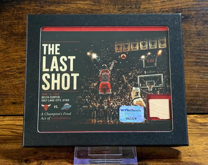May include: A black framed collectible display featuring a photograph of Michael Jordan taking The Last Shot in 1998. The print includes the text The Last Shot, June 14, 1998, Delta Center, Salt Lake City, Utah, a Bulls logo, and a holographic authenticity sticker.
