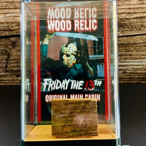 Op de afbeelding: Een doorzichtige vitrine met een houten relikwie uit de film "Friday the 13th". De vitrine heeft een afbeelding van Jason Voorhees en de tekst "MOOD RELIC WOOD RELIC" en "FRIDAY THE 13TH ORIGINAL MAIN CABIN."