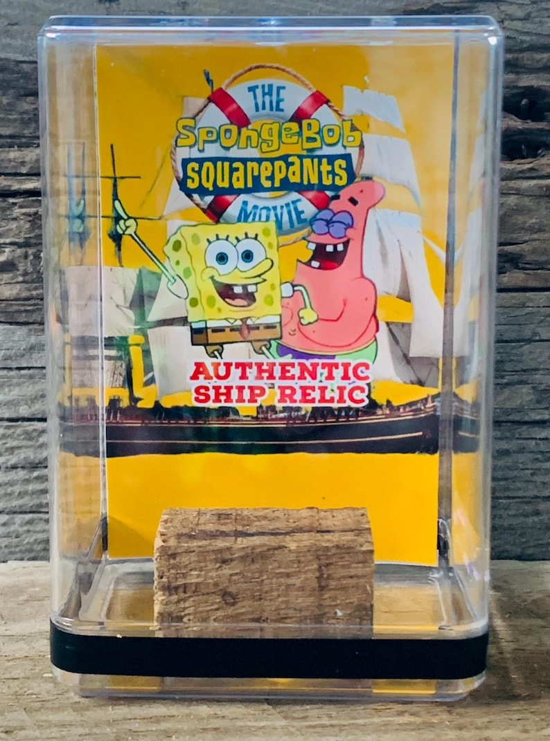 May include: A clear plastic display case with a yellow background featuring SpongeBob SquarePants and Patrick Star from the movie. The text "The SpongeBob SquarePants Movie" is printed on the background. A small piece of wood is displayed inside the case. The text "Authentic Ship Relic" is printed on the background.