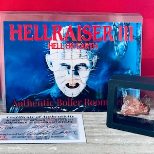 Może przedstawiać: Ramka z plakatem filmu grozy Hellraiser III: Hell on Earth. Plakat przedstawia zbliżenie postaci Pinheada, demonicznej postaci z twarzą pokrytą szpilkami. Plakat jest wystawiony obok małej czarnej gabloty, w której znajduje się mały, brązowy, kamienny przedmiot.