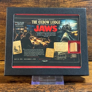 May include: A black framed display featuring a Jaws movie memorabilia card titled The Oxbow Lodge. The card includes text, a photo of the Orca boat, a shark, and a small piece of authentic wood from the lodge set within a red recessed square.