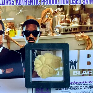 Puede incluir: Un trozo de piel amarilla enmarcado, posiblemente de un accesorio de pel&iacute;cula, se muestra frente a un p&oacute;ster con dos hombres con trajes negros y gafas de sol. El p&oacute;ster tiene las palabras "MIB BLACK" y "ARQUILLIANS AUTHENTIC PRODUCTION USED SKIN".
