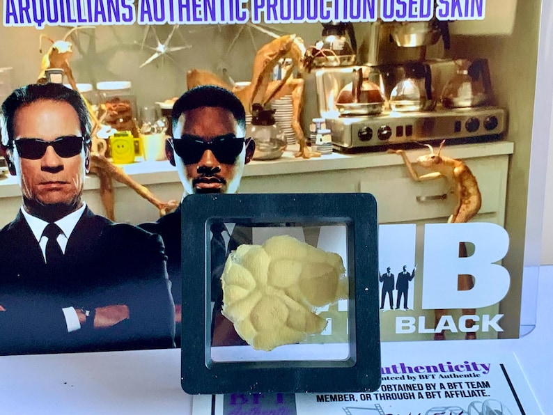 Puede incluir: Un trozo de piel amarilla enmarcado, posiblemente de un accesorio de pel&iacute;cula, se muestra frente a un p&oacute;ster con dos hombres con trajes negros y gafas de sol. El p&oacute;ster tiene las palabras "MIB BLACK" y "ARQUILLIANS AUTHENTIC PRODUCTION USED SKIN".
