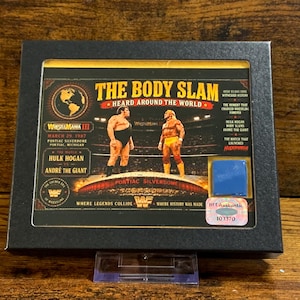 Könnte beinhalten: Gerahmte WrestleMania III-Erinnerungsstücke mit Hulk Hogan gegen Andre the Giant. Das Kunstwerk enthält den Titel der Veranstaltung, das Datum, den Ort und die Namen der Wrestler. Der Rahmen ist schwarz, und das Kunstwerk hat einen goldenen Rand.