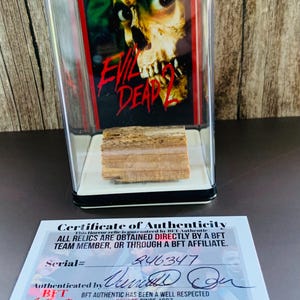 Op de afbeelding: Een doorzichtige vitrine met een stuk hout en een afbeelding uit de film "Evil Dead 2". De afbeelding toont een schedel met rode en gele tinten. Een certificaat van echtheid staat eronder.