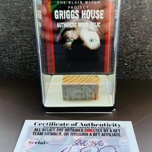 May include: A rectangular wooden relic from the Blair Witch Project's Griggs House, encased in a clear plastic display. The display includes a certificate of authenticity with a serial number and signature. The relic is a piece of wood.