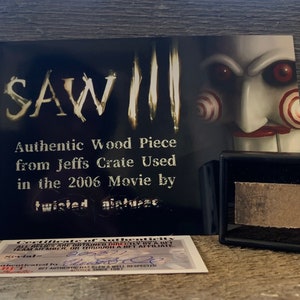 Puede incluir: Una tarjeta en blanco y negro con el texto "SAW!!!" en letras grandes. La tarjeta también tiene el texto "Authentic Wood Piece from Jeffs Crate Used in the 2006 Movie by twistedpictures." Una pequeña caja negra con un trozo de madera en su interior se encuentra en la esquina inferior derecha de la imagen. La tarjeta también tiene un certificado de autenticidad con el número de serie 02892.