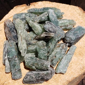 May include: A collection of rough, raw kyanite crystals. The crystals are a mix of blue-green and gray, with a natural, textured appearance. The kyanite pieces are piled together, showcasing their unique shapes and mineral formations.
