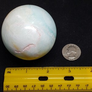 May include: A large, light blue, green, and white sphere with pink veins. The sphere is about 2 inches in diameter and is sitting on a black surface. A ruler and a US quarter are in the image for scale.