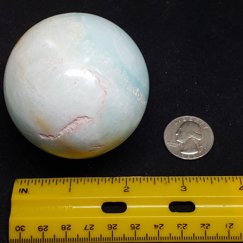 May include: A large, light blue, green, and white sphere with pink veins. The sphere is about 2 inches in diameter and is sitting on a black surface. A ruler and a US quarter are in the image for scale.