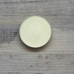 Könnte beinhalten: Ein runder Behälter mit silbernem Rand, gefüllt mit einer hellgelben Substanz, möglicherweise ein Kosmetikprodukt. Der Behälter befindet sich auf einer verwitterten, grauen Holzoberfläche.