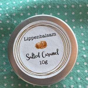 Könnte beinhalten: Kleine, runde, silberne Dose Lippenbalsam mit weißem Etikett. Das Etikett zeigt die Wörter "Lippenbalsam" und "Salted Caramel" in handschriftlicher Schrift, zusammen mit einer Karamellgrafik und "10g". Die Dose liegt auf einer türkisfarbenen, gepunkteten Oberfläche.
