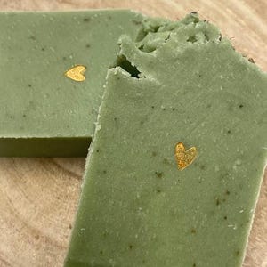 Könnte beinhalten: Zwei grüne Seifenstücke mit einem goldenen Herzdesign. Die Seife hat eine strukturierte Oberfläche und sichtbare Flecken. Ein Stück ist teilweise zerbrochen und zeigt eine raue Kante. Die Seife ist hellgrün.
