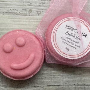 Könnte beinhalten: Zwei rosa Shampoo-Bars. Einer hat die Form eines Smiley. Der andere ist rund und in einem rosa Netzbeutel verpackt. Auf dem Etikett steht "Shampoo Bar English Rose" und die Inhaltsstoffe sind aufgelistet. Jede Bar wiegt 45g.
