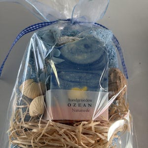 Könnte beinhalten: Ein Geschenkset, verpackt in durchsichtigem Zellophan, mit einem blau-weißen Seifenstück mit dem Text "handgesiedete OZEAN Naturseife", einem blauen Waschlappen, dekorativen Muscheln und Bast. Das Set ist mit einem blauen Band und einem transparenten blauen Blumenband gebunden.