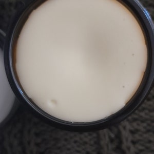Op de afbeelding: Een zwarte pot met een wit deksel met een witte cr&egrave;me of lotion.