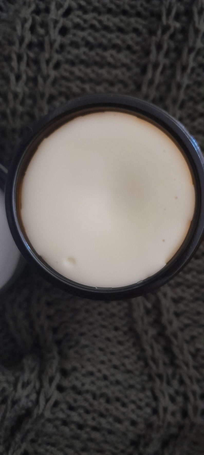 Op de afbeelding: Een zwarte pot met een wit deksel met een witte cr&egrave;me of lotion.