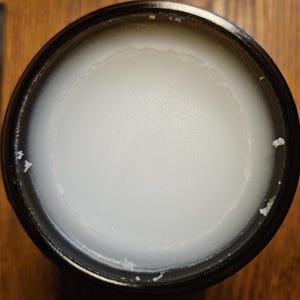 Puede incluir: Un recipiente negro lleno de una sustancia blanca y cremosa. El producto tiene una superficie lisa y ligeramente texturizada. El recipiente es redondo con un borde oscuro.