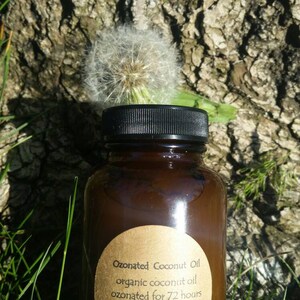 May include: A brown glass bottle with a black lid and a gold label that reads "Ozonated Coconut Oil organic coconut oil ozonated for 72 hours OzonedOrganics.com". The bottle is sitting on a bed of green grass.
