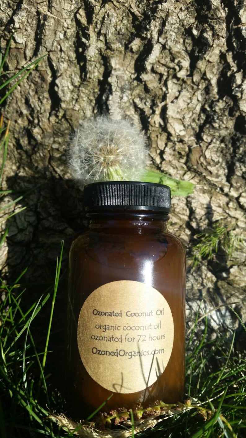 May include: A brown glass bottle with a black lid and a gold label that reads "Ozonated Coconut Oil organic coconut oil ozonated for 72 hours OzonedOrganics.com". The bottle is sitting on a bed of green grass.