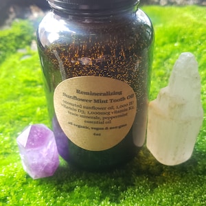May include: A brown glass bottle with a label that reads "Remineralizing Sunflower Mint Tooth Oil" and lists ingredients including ozonated sunflower oil, vitamin D3, vitamin K2, trace minerals, peppermint essential oil. The label also states that the product is all organic, vegan, and non-GMO. The bottle is sitting on a bed of green moss with two crystals, one purple and one clear, on either side of the bottle.